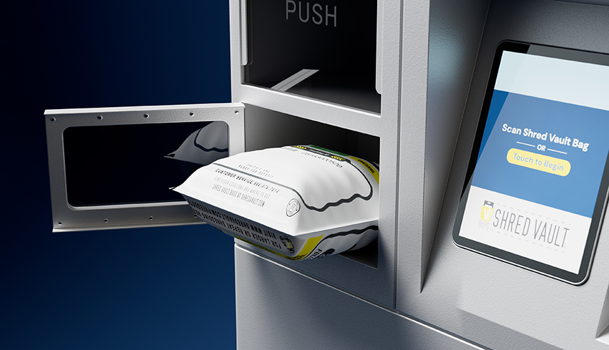 A close-up of a secure shredding kiosk. A hand-sized white Shred Vault bag is partially inserted into a rectangular metal deposit slot labeled “PUSH.” To the right, a digital screen displays instructions that read “Deposit Shred Vault Bag” with a visual guide showing how to insert the bag. The machine has a sleek, metallic finish, and the background is a dark blue gradient.